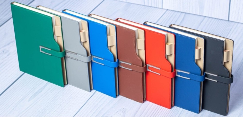 Seven colorful, closed, leather-bound planners with metal clasps (Green, Grey, Blue, Brown, Red, Navy, Charcoal).